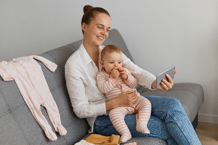 A smiling babysitter with a baby on a couch, holding a phone, ready to expose the aunt.