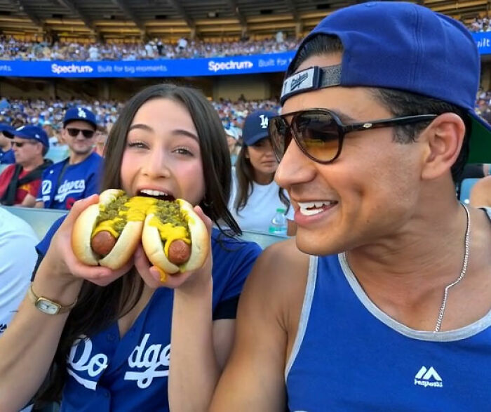 Mario Lopez and his niece smiling while eating hot dogs at a baseball game.