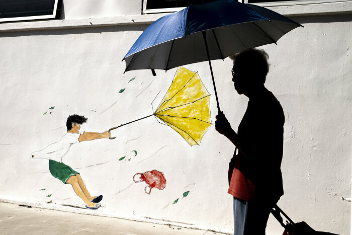 A person with a blue umbrella walks past a street photography mural of a boy holding a yellow umbrella in art.