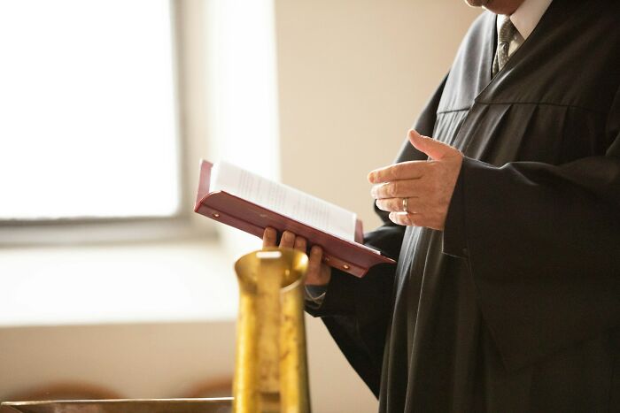 A person in a black robe holding a book and gesturing with their hand during a wedding ceremony.