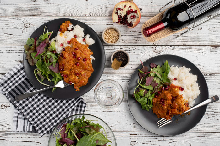 A top-down view of two plates of food, salad, rice, and a red sauce with pomegranates, with a bottle of wine nearby. This image represents the bestie pulls away after supporting woman through divorce. A top-down view of two plates of food, salad, rice, and a red sauce with pomegranates, with a bottle of wine nearby. This image represents the bestie pulls away after supporting woman through divorce.