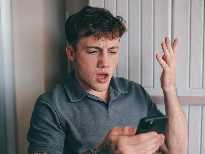 A man with curly hair and a grey polo shirt looks confused and gestures with one hand while holding a phone in the other. This represents a bestie pulls away after supporting woman through divorce. A man with curly hair and a grey polo shirt looks confused and gestures with one hand while holding a phone in the other. This represents a bestie pulls away after supporting woman through divorce.