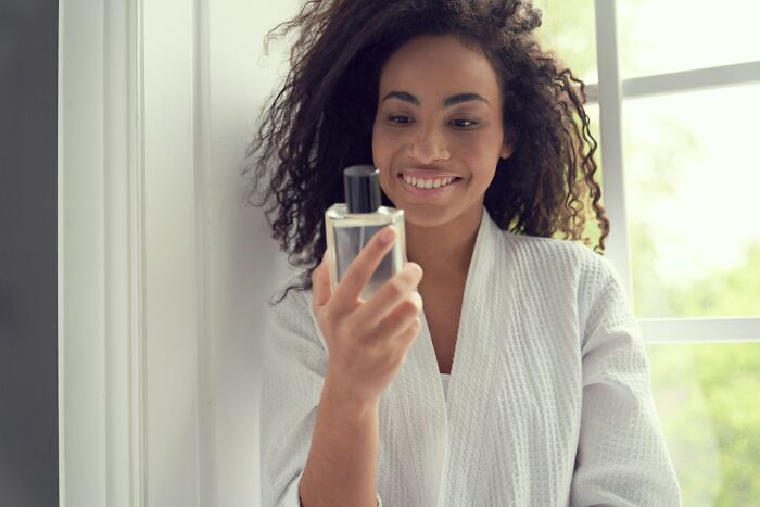 A smiling woman in a bathrobe holding a perfume bottle, enjoying self-care while living alone and being single.