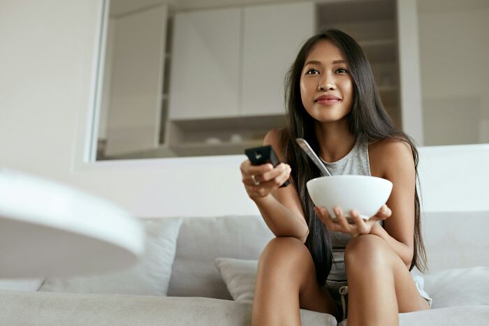 A young woman enjoying a snack and watching TV, embodying the relaxed lifestyle of living alone and being single.
