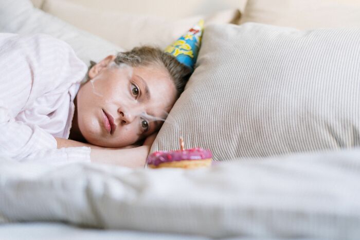 A woman wearing a party hat, looking sad next to a donut with a candle, representing feelings while living alone.
