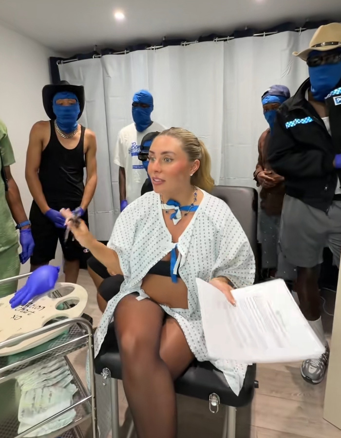 Bonnie Blue in a hospital gown holding papers, seated among several men in blue balaclavas during a stunt. Bonnie Blue in a hospital gown holding papers, seated among several men in blue balaclavas during a stunt.