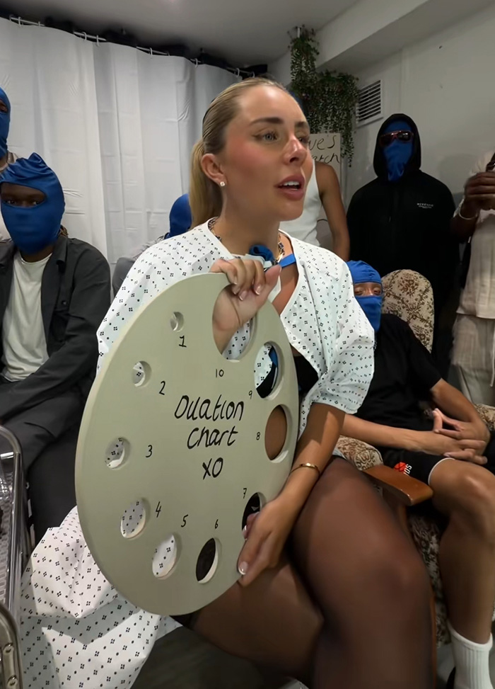 Bonnie Blue in a hospital gown holding a circular chart, surrounded by people in blue balaclavas during a stunt. Bonnie Blue in a hospital gown holding a circular chart, surrounded by people in blue balaclavas during a stunt.