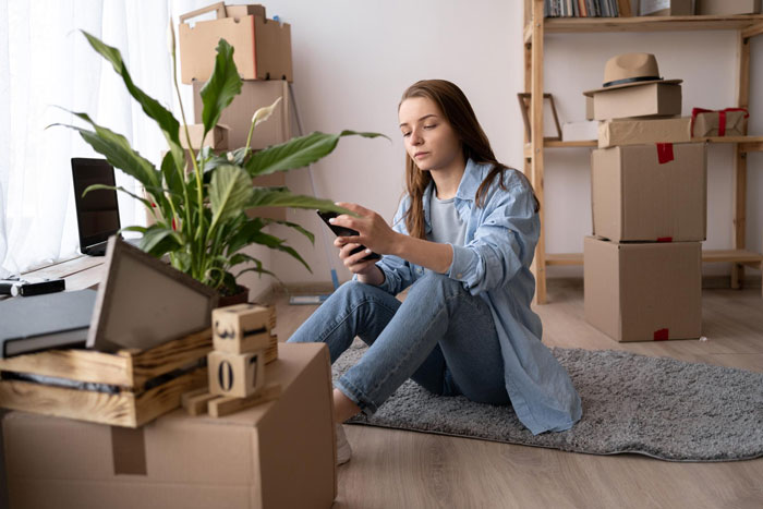 Ex-girlfriend sends receipts: A young woman sits among moving boxes, looking at her phone. Ex-girlfriend sends receipts: A young woman sits among moving boxes, looking at her phone.