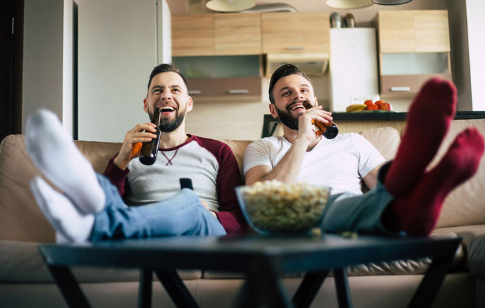 Two friends relaxing on a couch, enjoying beer and popcorn, while watching TV, unaware of a man playing friends against each other. Two friends relaxing on a couch, enjoying beer and popcorn, while watching TV, unaware of a man playing friends against each other.