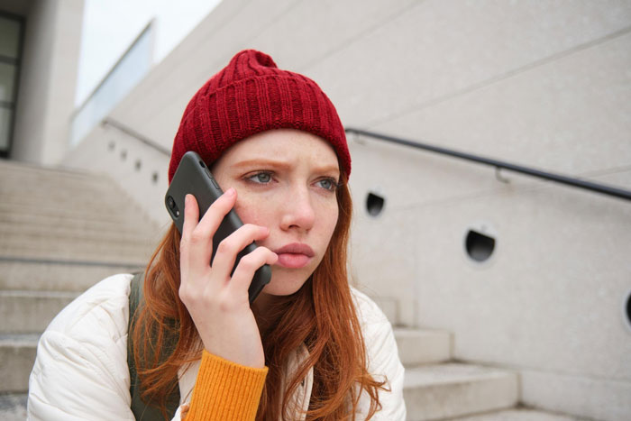 A red-haired woman with a red beanie talking on a phone, relating to a man playing his friends against each other. A red-haired woman with a red beanie talking on a phone, relating to a man playing his friends against each other.