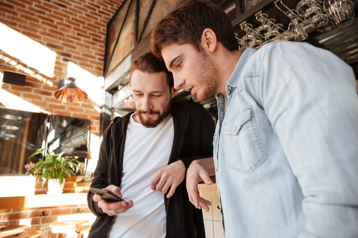Two men looking at a phone, one with a beard and black hoodie, the other in a denim shirt. Two men looking at a phone, one with a beard and black hoodie, the other in a denim shirt.