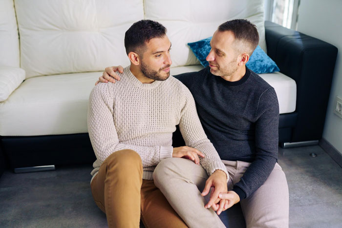 Two men sitting on the floor, holding hands, depicting a relationship unraveling due to a conniving boyfriend. Two men sitting on the floor, holding hands, depicting a relationship unraveling due to a conniving boyfriend.