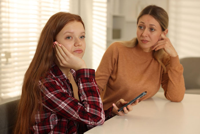 A woman looking upset with her hand on her cheek, holding a phone, as her mother looks at her worriedly in the background. Illustrates the impact of a toxic sister. A woman looking upset with her hand on her cheek, holding a phone, as her mother looks at her worriedly in the background. Illustrates the impact of a toxic sister.