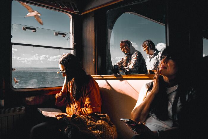 Passengers inside a train car, one reading, others looking out at the sea and sky, an award-winning street photography.