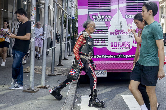 An elderly person in elaborate punk attire with platform boots walks past a pink ice cream truck, a vibrant street photography scene.
