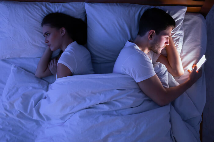 A couple in bed, turning their backs to each other, the man looking at his phone, representing relationship red flags and mental health.