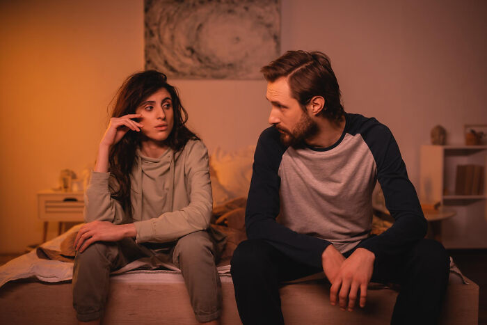 A couple sits on a bed, looking upset, possibly discussing reasons for an affair.