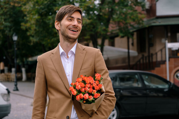 A smiling man in a brown jacket holds a bouquet of orange flowers, possibly a gift for affair partners.
