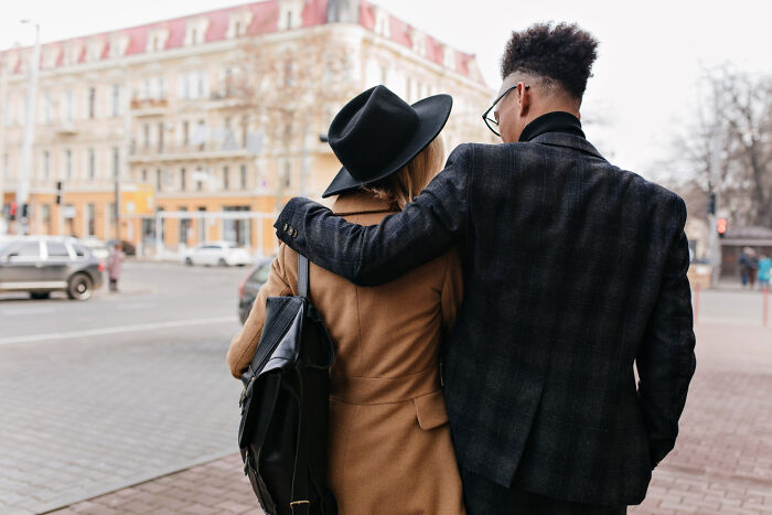Rear view of a couple with arms around each other, walking down a city street, representing affair partners.