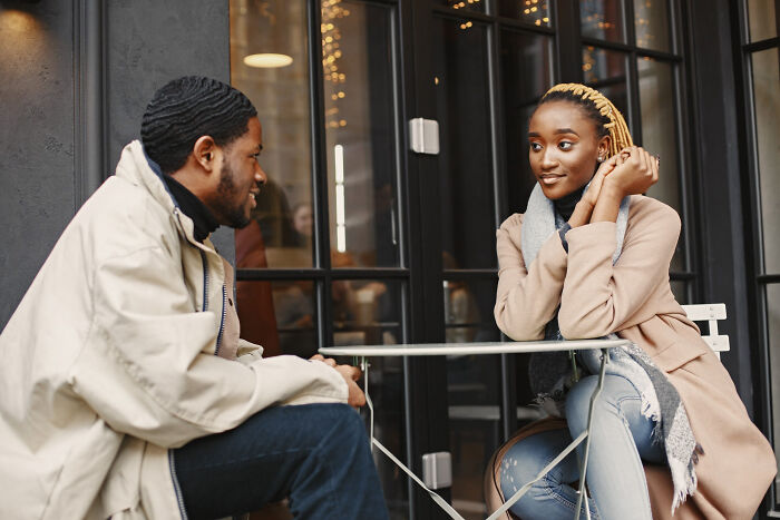 A man and a woman sitting across from each other at an outdoor cafe table, discussing reasons affair partners have.