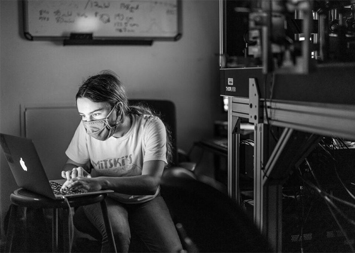 A scientist wearing a mask working on a laptop, surrounded by lab equipment. Scientists discover new color Olo.