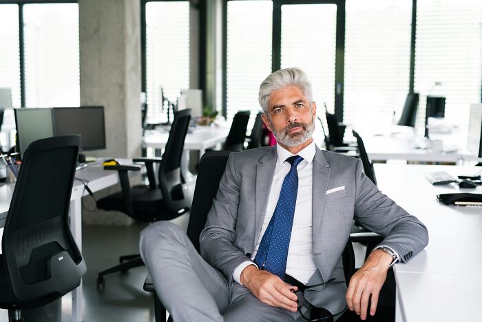 A stylish grey-haired man in a suit sits confidently in an office, showcasing insider knowledge of corporate industries.