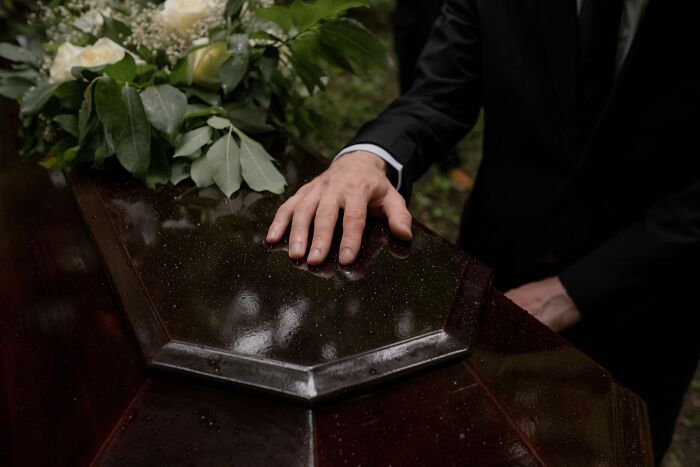 A man in a black suit places his hand on a dark wooden coffin, representing insider knowledge of funeral industries.