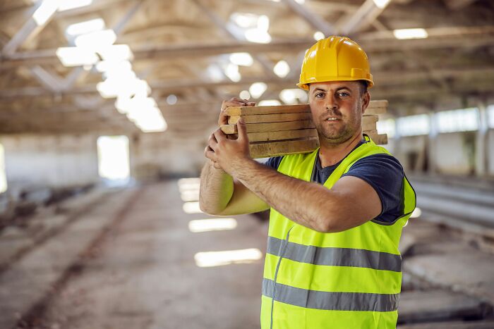 A construction worker in a yellow hard hat and safety vest carries lumber on his shoulder, sharing insider knowledge.