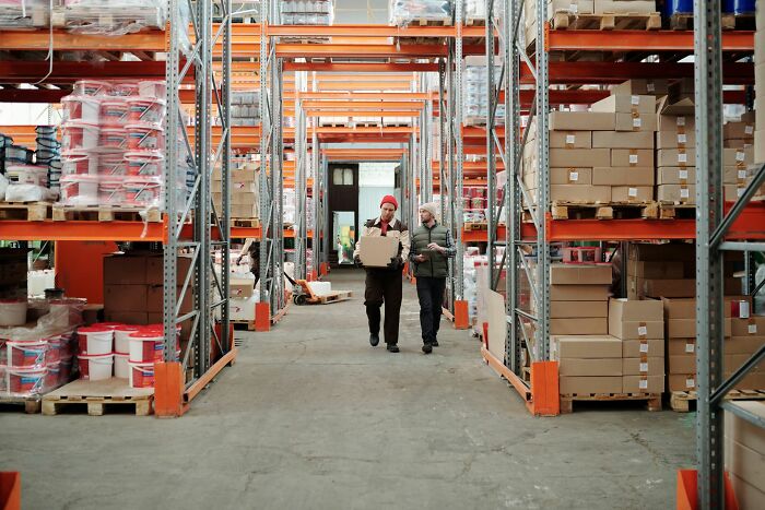 Two workers carrying a box down an aisle of a large warehouse, showing a company in operation.
