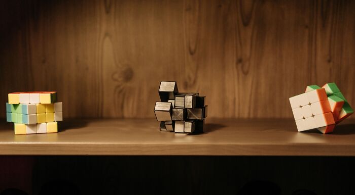 Three puzzle cubes on a wooden shelf, a colorful 2x2, a reflective mirror cube, and a partially solved 3x3 Rubik's cube, for music puzzles.