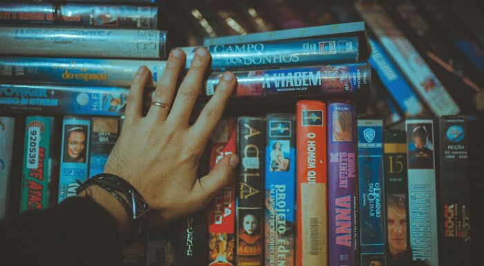Hand reaching for a VHS tape on a shelf filled with various movies, testing your movie IQ with rebus challenges.