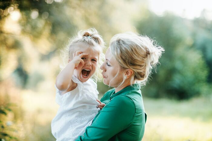 A mother holding a crying toddler outdoors, with the child making a distressed expression. Teachers often face such situations.