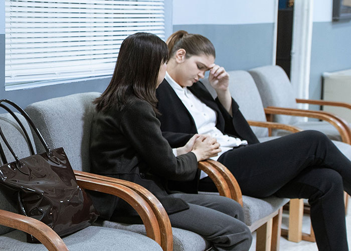 Two women sitting in a waiting room, one comforting the other, who appears distressed, likely due to a cheating soulmate.