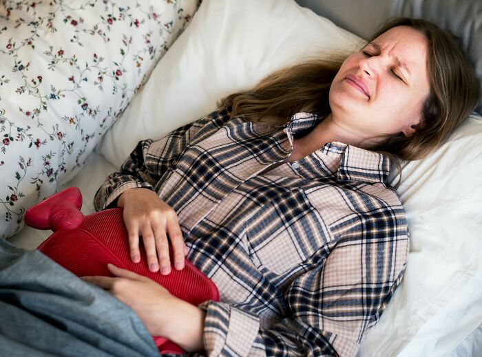 A woman lies in bed, grimacing in pain and holding a hot water bottle, reflecting on unhinged things doctors say.