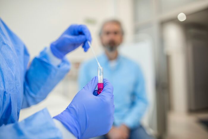 A medical professional in blue gloves holds a test tube with a swab, with a patient in the background; reflecting unhinged doctors.