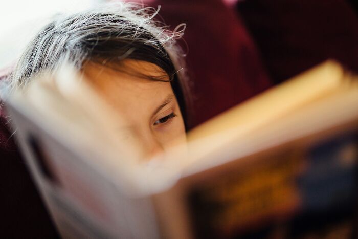 A young girl with dark hair reading a book, her eyes focused, lost in a story that might involve strange experiences.
