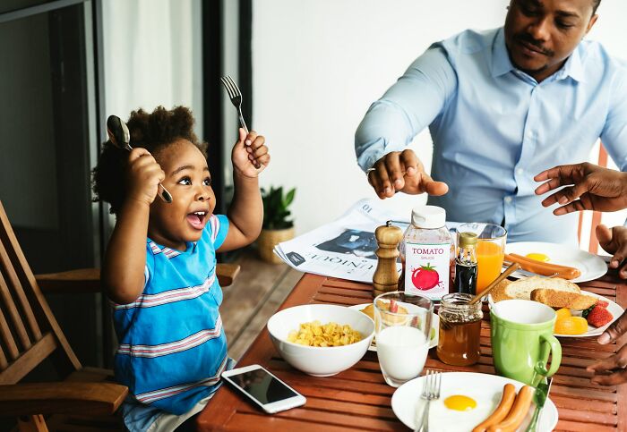 A happy child holding utensils excitedly at a breakfast table with an adult nearby, contrasting with a horrible parent.