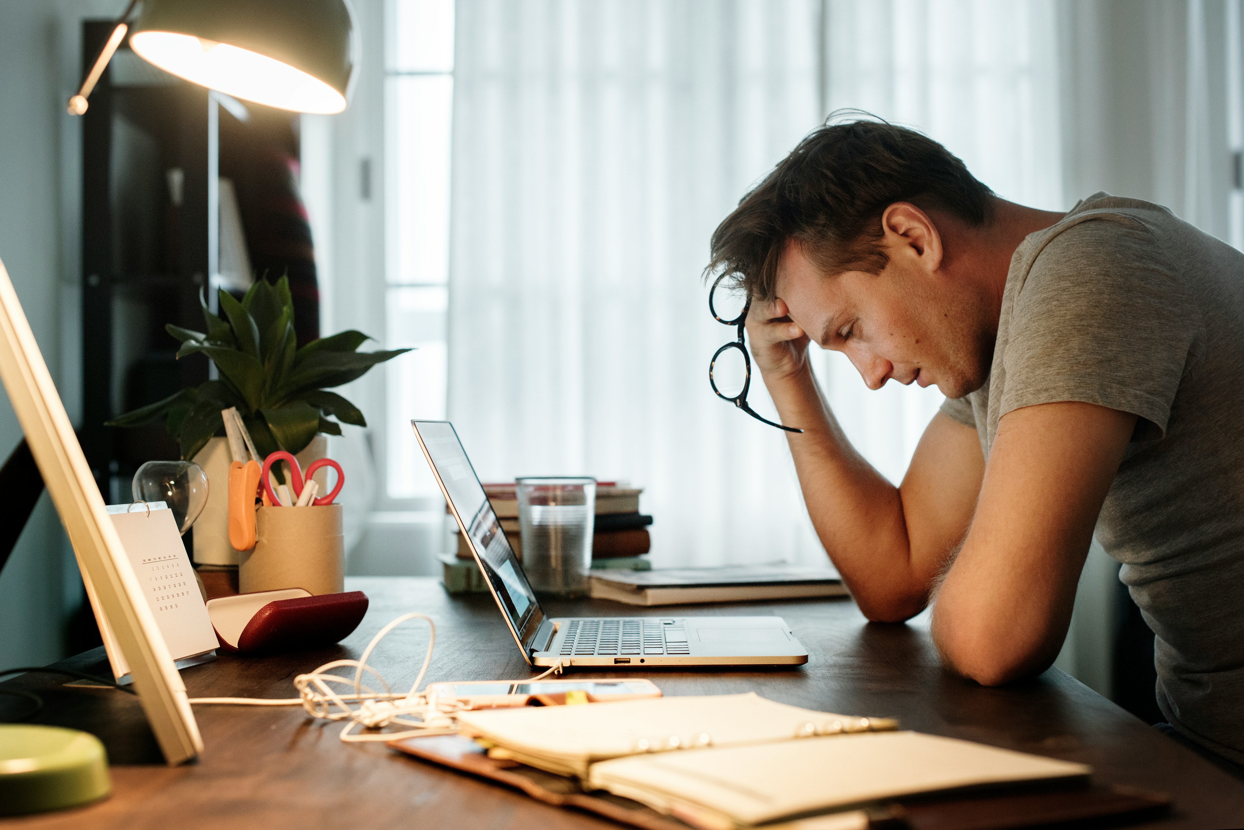 A man, frustrated and stressed, holds his glasses while looking at his fancy laptop on a cluttered desk. A man, frustrated and stressed, holds his glasses while looking at his fancy laptop on a cluttered desk.