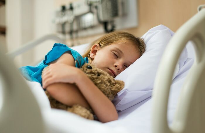A young girl sleeping in a hospital bed, hugging a teddy bear, illustrating a patient receiving diagnoses.