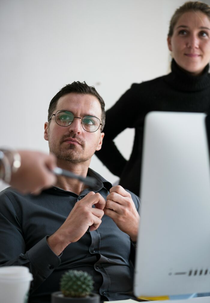 Man with glasses buttoning his shirt while a woman stands behind him, capturing sick burns of everyday life.