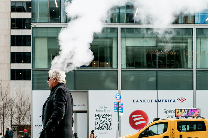 A man with white hair and sunglasses walks past a steamy street vent in front of a modern building, a scene of street photography.