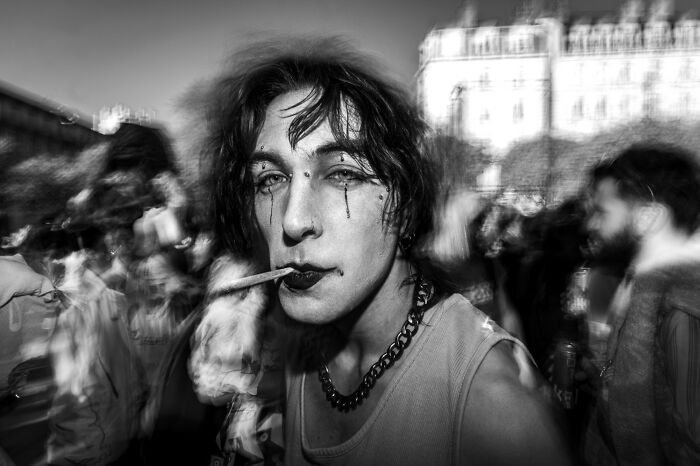 A person with dark eye makeup and a cigarette in their mouth, a street photo from the Macadam Awards.
