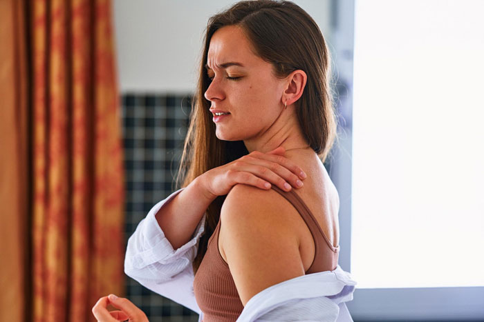 A woman with long brown hair, wearing a brown top and white shirt, holds her shoulder in pain, representing a person with a disability.