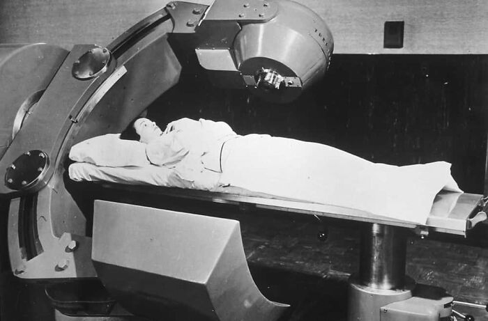 A patient on a table, receiving a terrifying medical treatment with a large, rotating machine in the past.