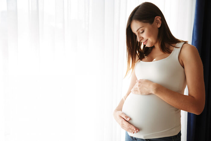 A pregnant woman gently cradling her baby bump by a window, representing patient care.