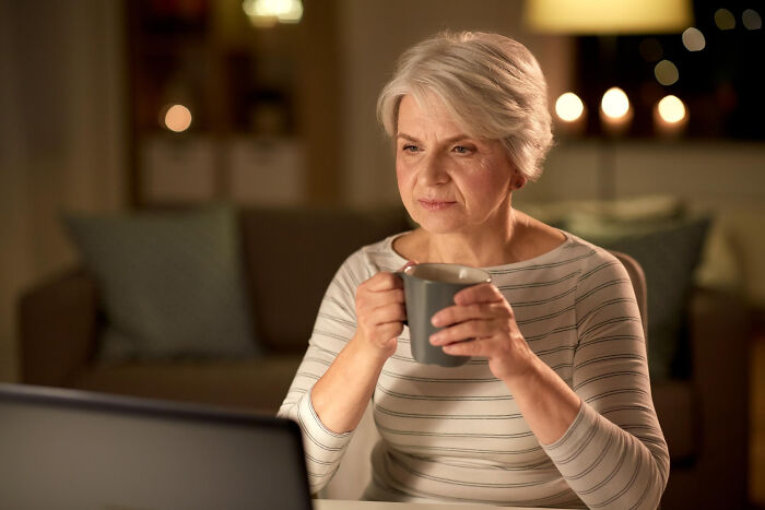 An older woman with gray hair sips from a mug while looking at a laptop, pondering stupid things patients have said.