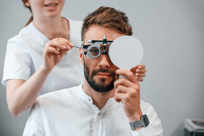 Male patient having an eye exam with a phoropter, covering one eye while the doctor adjusts the lens.