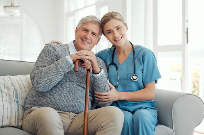 A happy female doctor and a smiling elderly male patient with a cane, sitting together on a sofa, highlighting patient care.