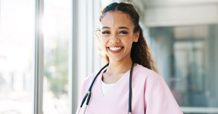 Smiling young female doctor in pink scrubs and a stethoscope standing in a bright hospital hallway.