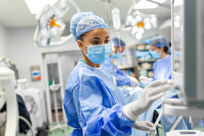 Female doctor in scrubs, mask, and surgical cap looking at a medical monitor in an operating room with other doctors.